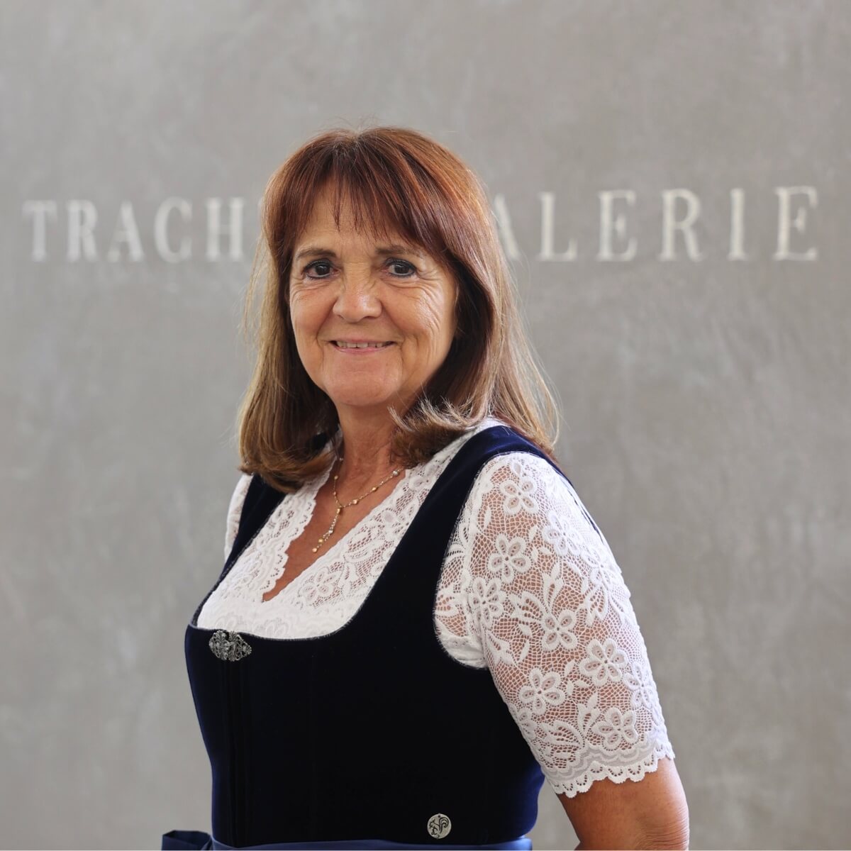 Renate, eine freundliche Mitarbeiterin der Trachtengalerie, steht in einem eleganten Dirndl vor einem Wandhintergrund mit dem Schriftzug 'Trachtengalerie'.