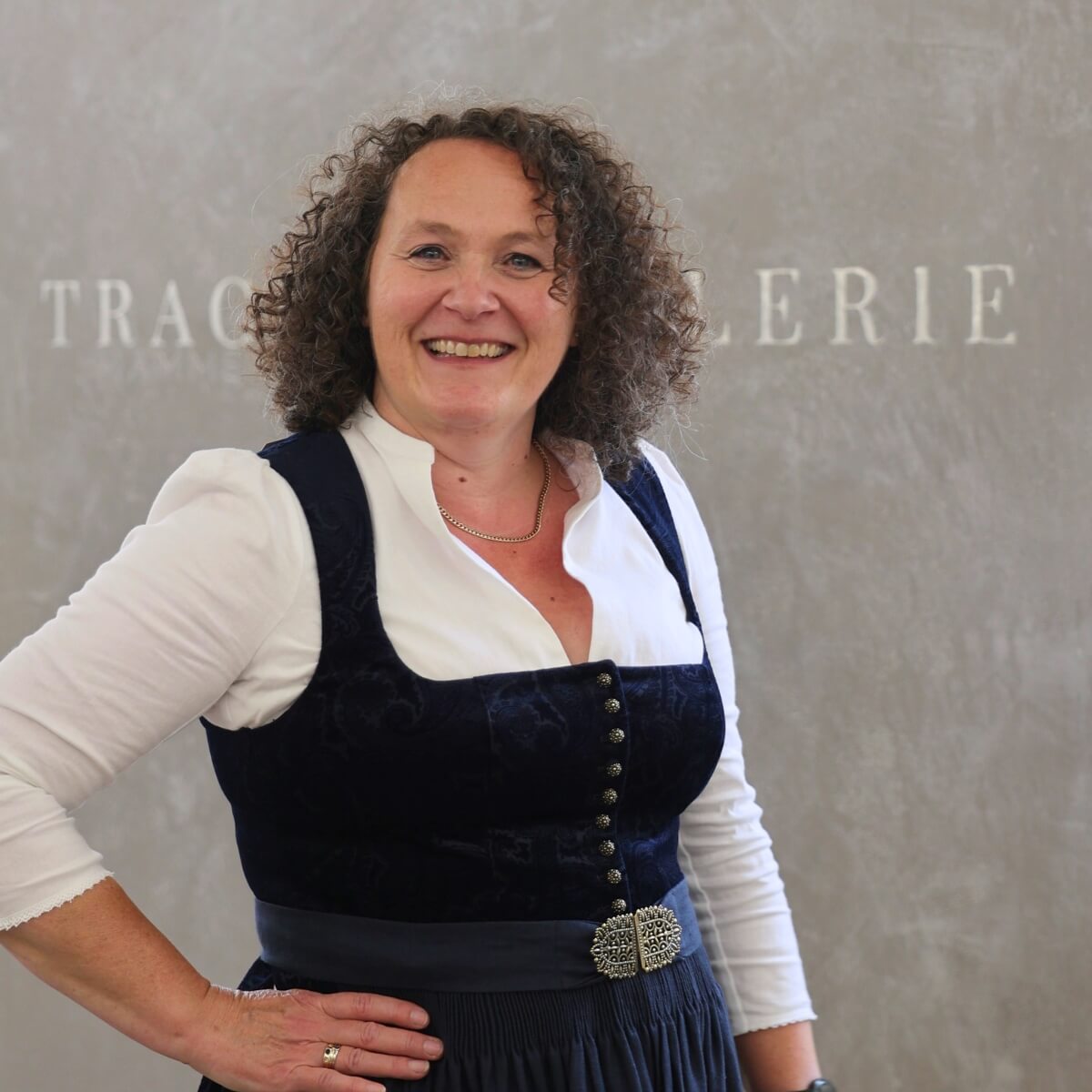 Gesine, eine erfahrene Dirndlfachverkäuferin der Trachtengalerie, steht in einem schönen Dirndl vor einem Wandhintergrund mit dem Schriftzug 'Trachtengalerie'.