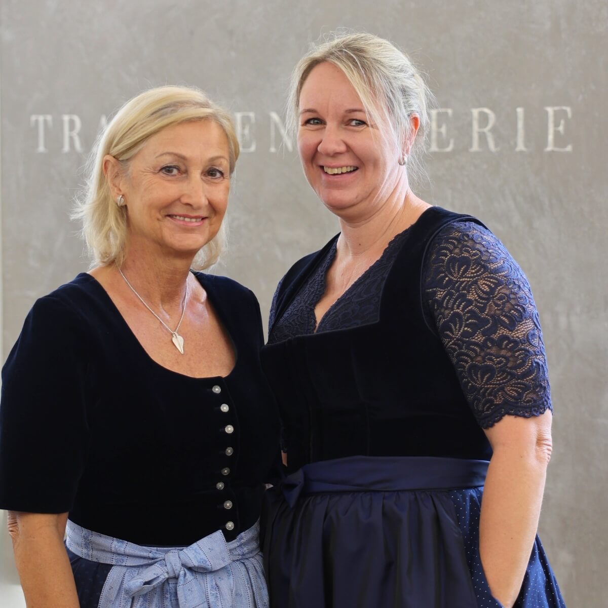 Erna und Michaela, zwei freundliche Mitarbeiterinnen der Trachtengalerie, stehen vor einem Wandhintergrund mit dem Schriftzug 'Trachtengalerie'. Beide tragen traditionelle Dirndl.