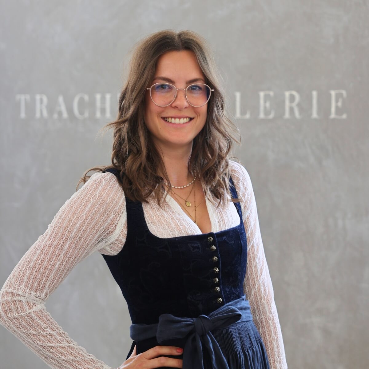 Dani, eine freundliche Aushilfe der Trachtengalerie, steht vor einem Wandhintergrund mit dem Schriftzug 'Trachtengalerie'. Sie trägt ein traditionelles Dirndl.