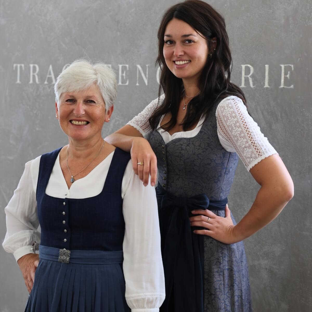 Beate und Eva, zwei freundliche Mitarbeiterinnen der Trachtengalerie, stehen vor einem Wandhintergrund mit dem Schriftzug 'Trachtengalerie'. Beide tragen traditionelle Dirndl.