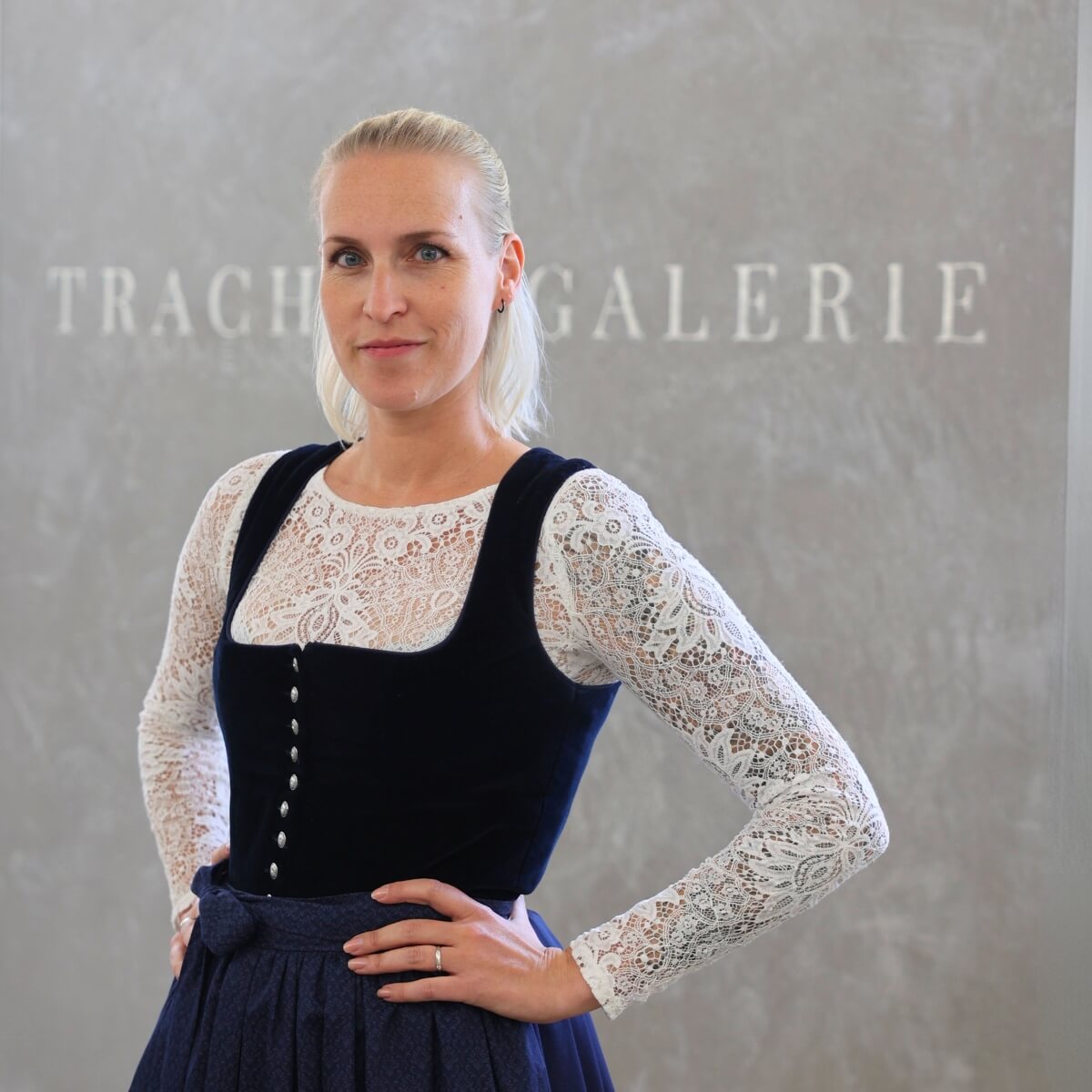 Vanessa, eine freundliche Aushilfe der Trachtengalerie, steht vor einem Wandhintergrund mit dem Schriftzug 'Trachtengalerie'. Sie trägt ein traditionelles Dirndl und lächelt herzlich.