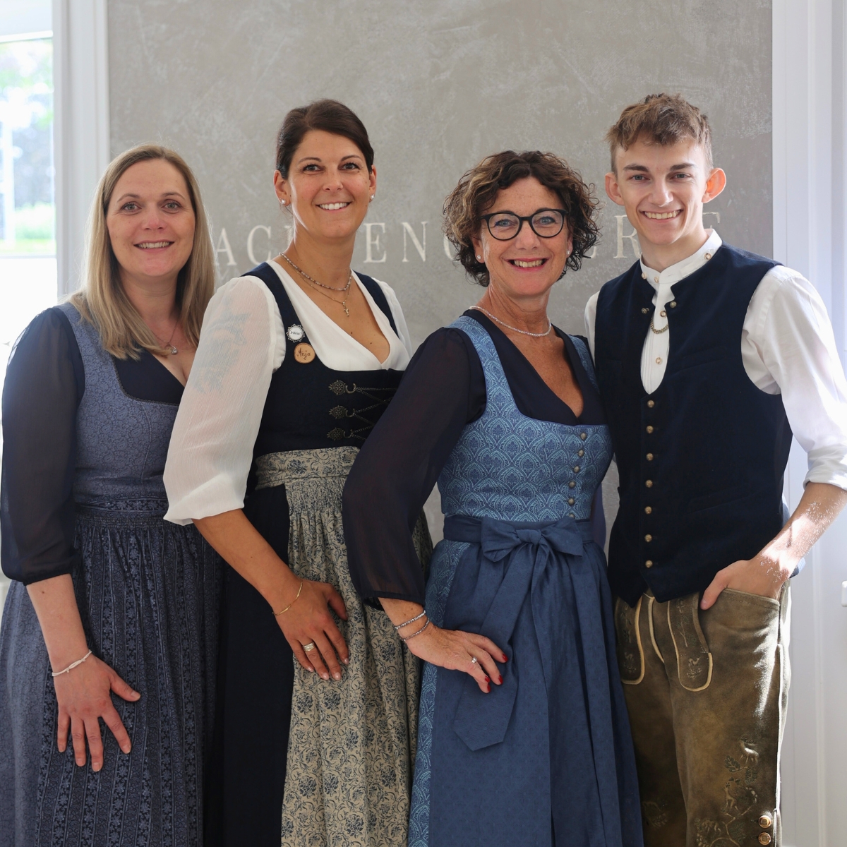 Petra, Anja, Geli und Finn-Luca, Mitarbeiter der Herrenabteilung der Trachtengalerie, stehen gemeinsam in traditioneller Tracht vor einem Wandhintergrund mit dem Schriftzug 'Trachtengalerie'.